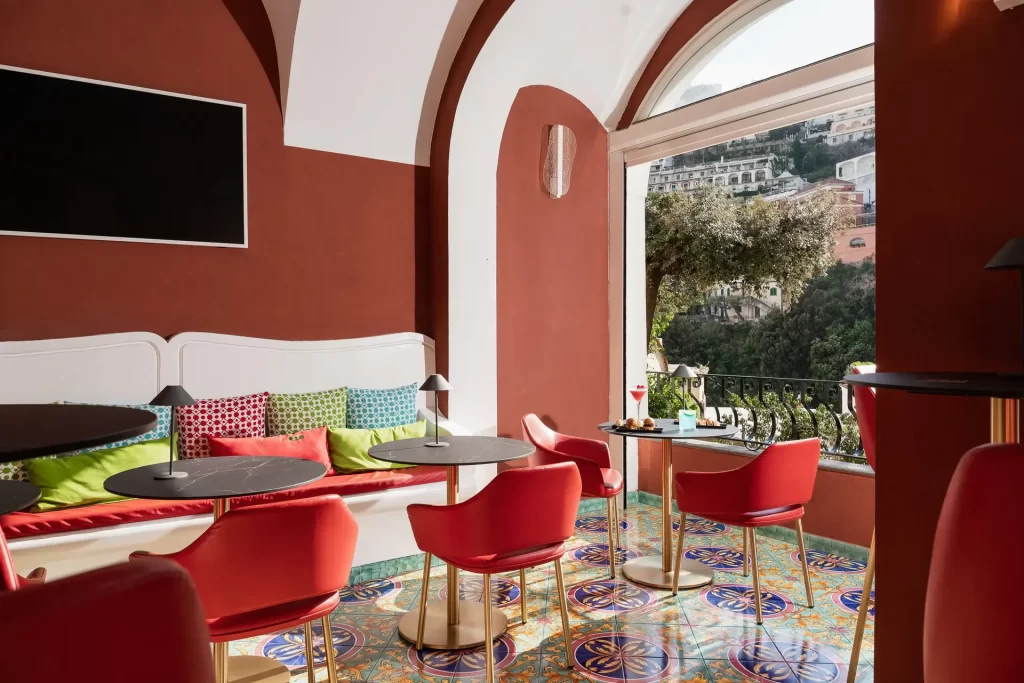 Sala interna elegante con sedie rosse, tavoli moderni, pavimento colorato e vista su un paesaggio mediterraneo fuori dalla finestra.