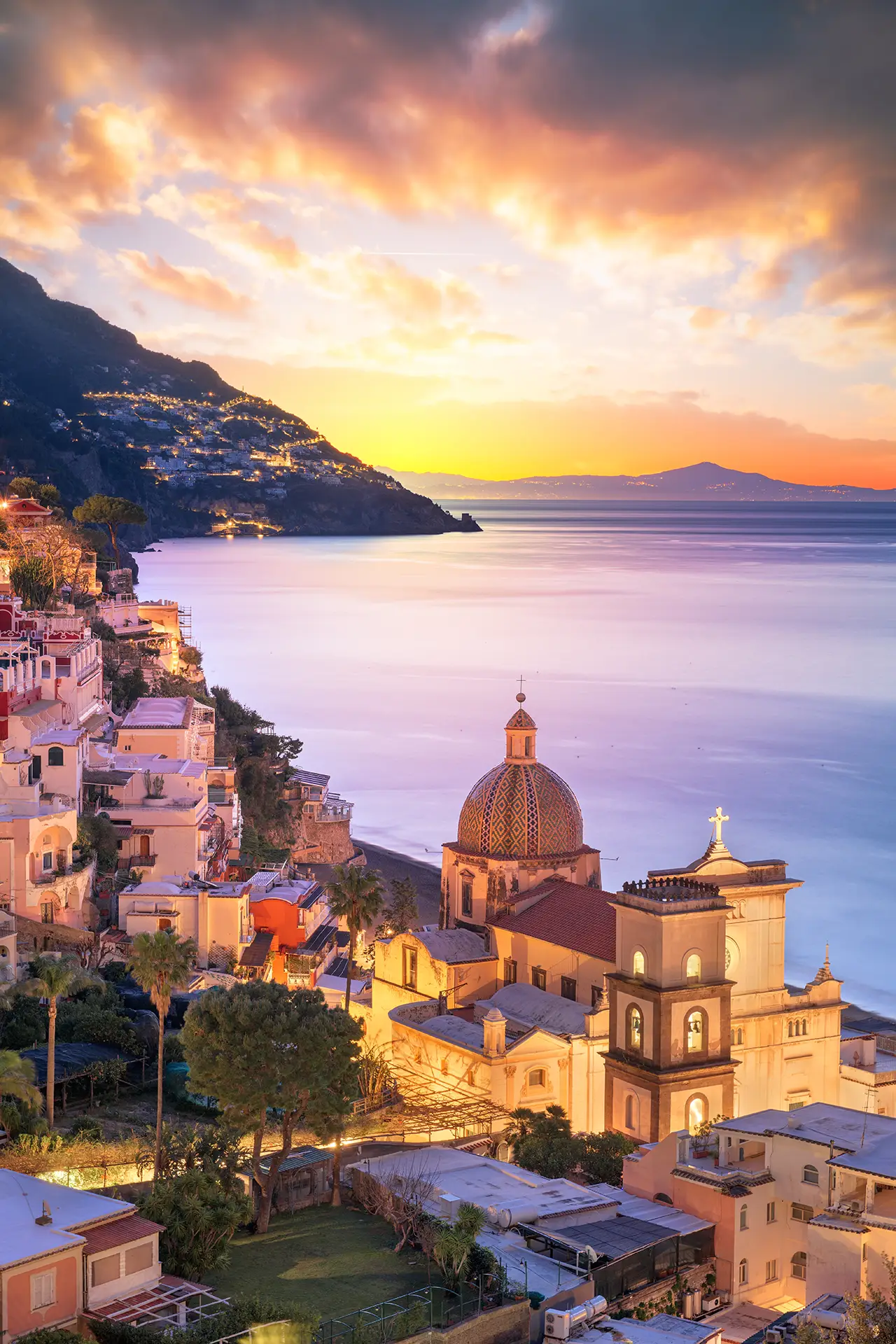 A scenic view of a coastal town at sunset with colorful buildings and a dome-shaped church.